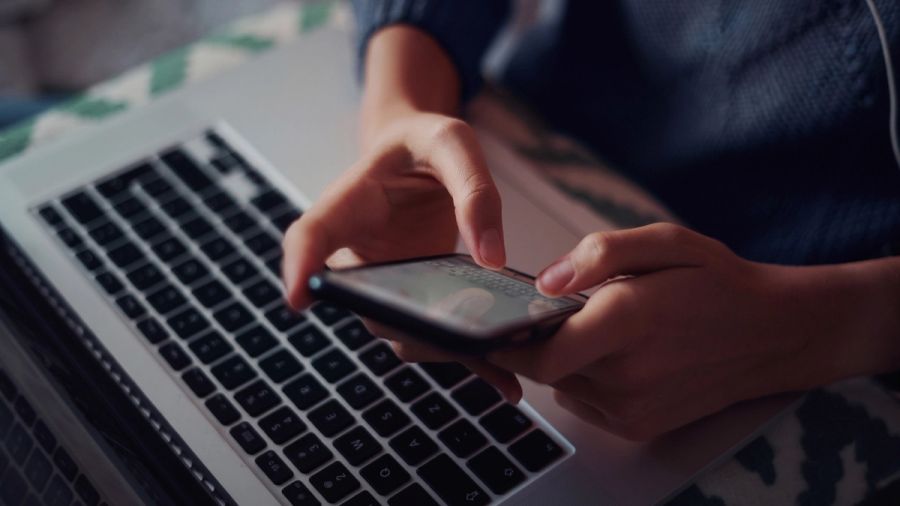 Hands using a smartphone above a laptop keyboard.