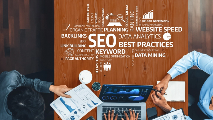 Overhead view of two people working at a desk covered with SEO keywords and icons, with a laptop displaying data analytics.