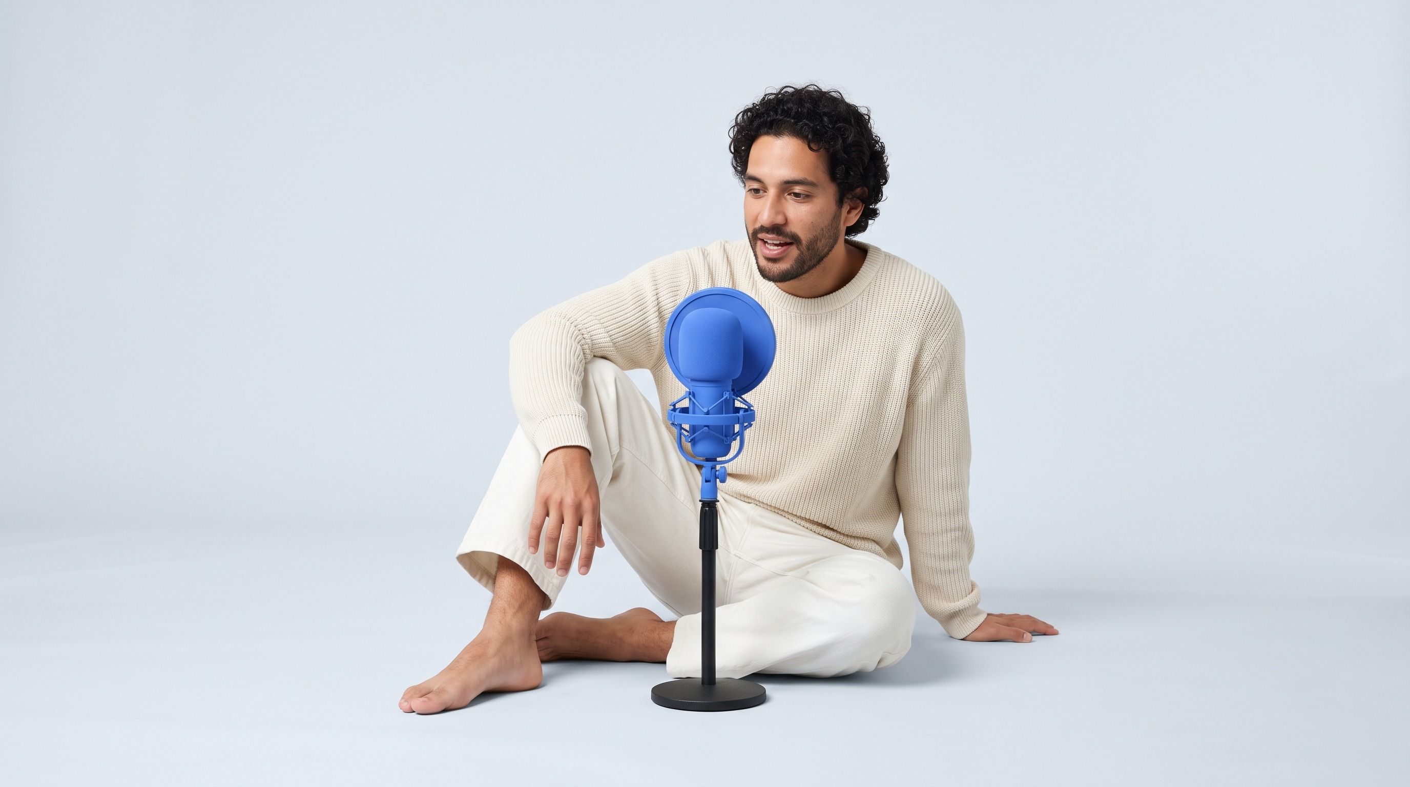 A man with curly hair sits on the floor, speaking into a blue microphone.