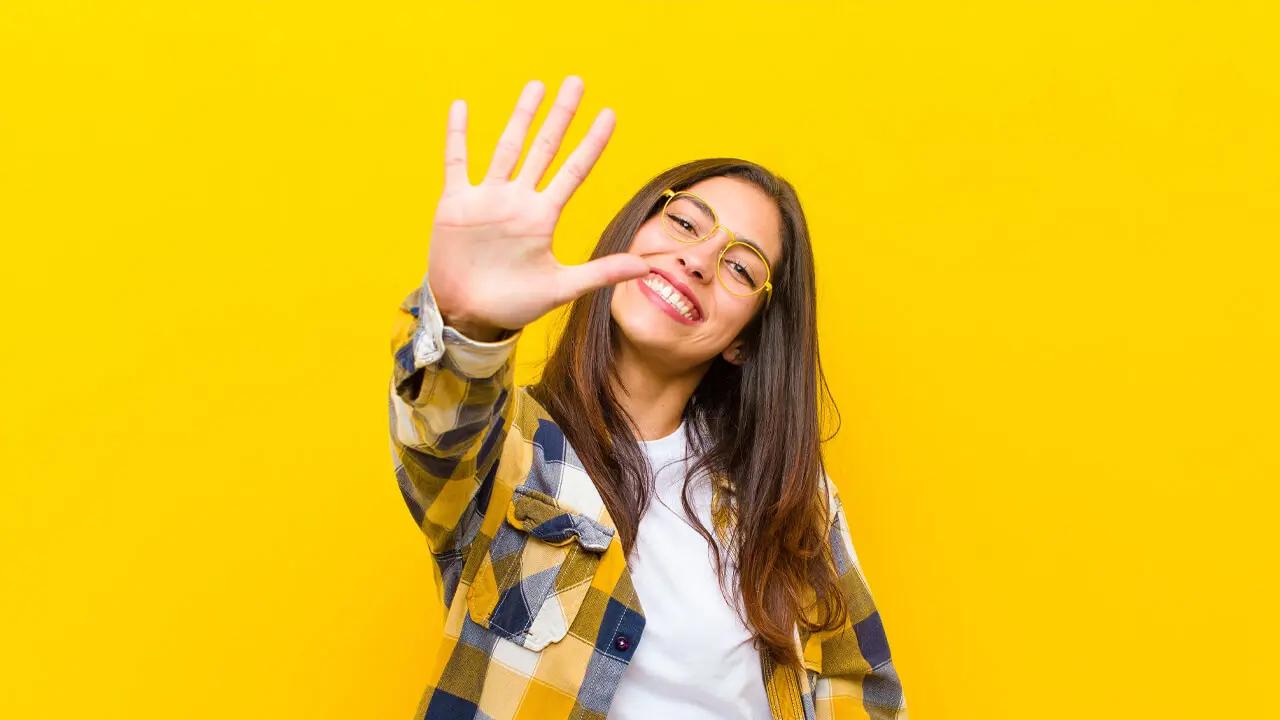 A smiling woman in glasses and a plaid shirt holds up her hand, showing five fingers.