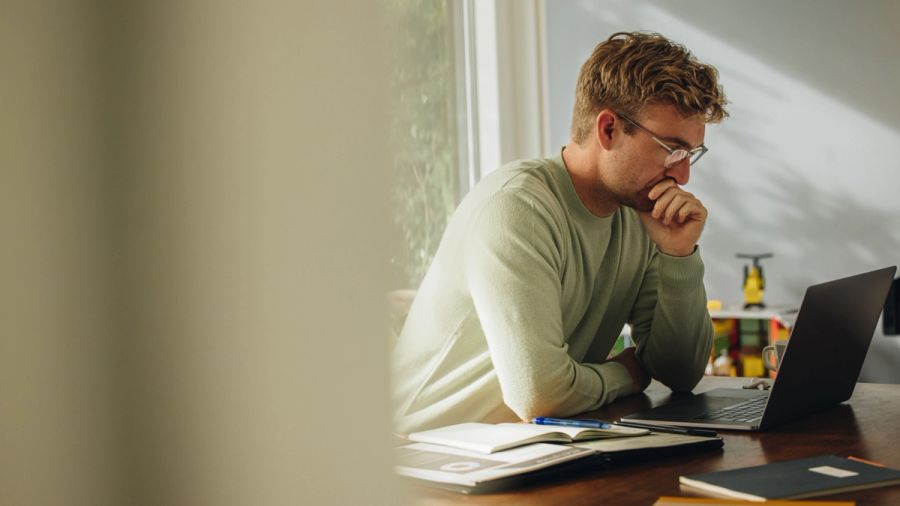 A focused man in glasses looks at a laptop with his hand on his chin.