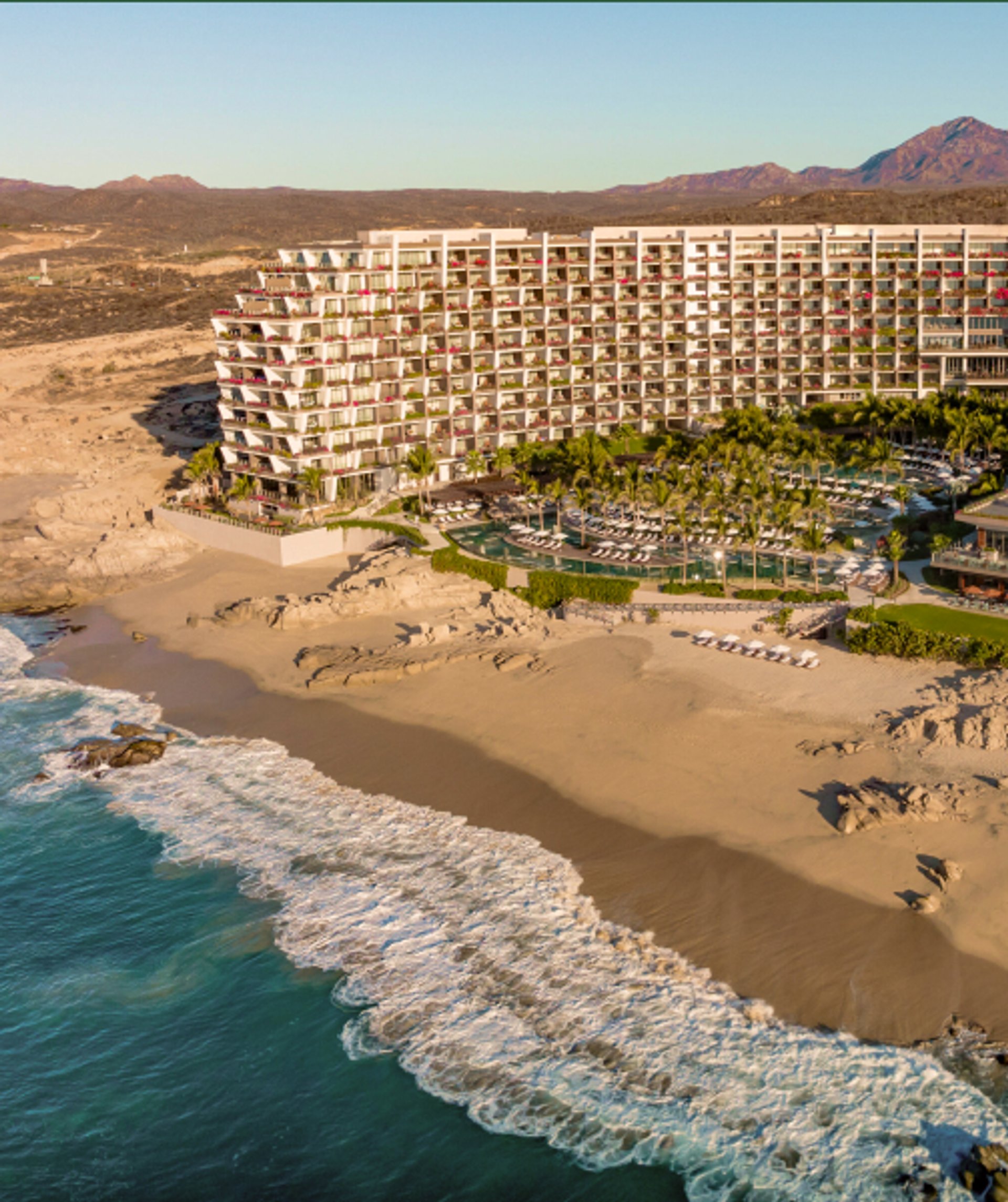 Aerial view of a large white resort complex on a sandy beach with pools, palm trees, and ocean waves, set against arid mountains.