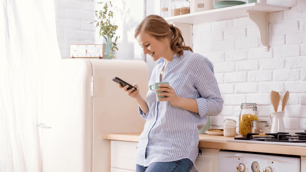A smiling woman holding a mug and looking at her smartphone in a bright kitchen.