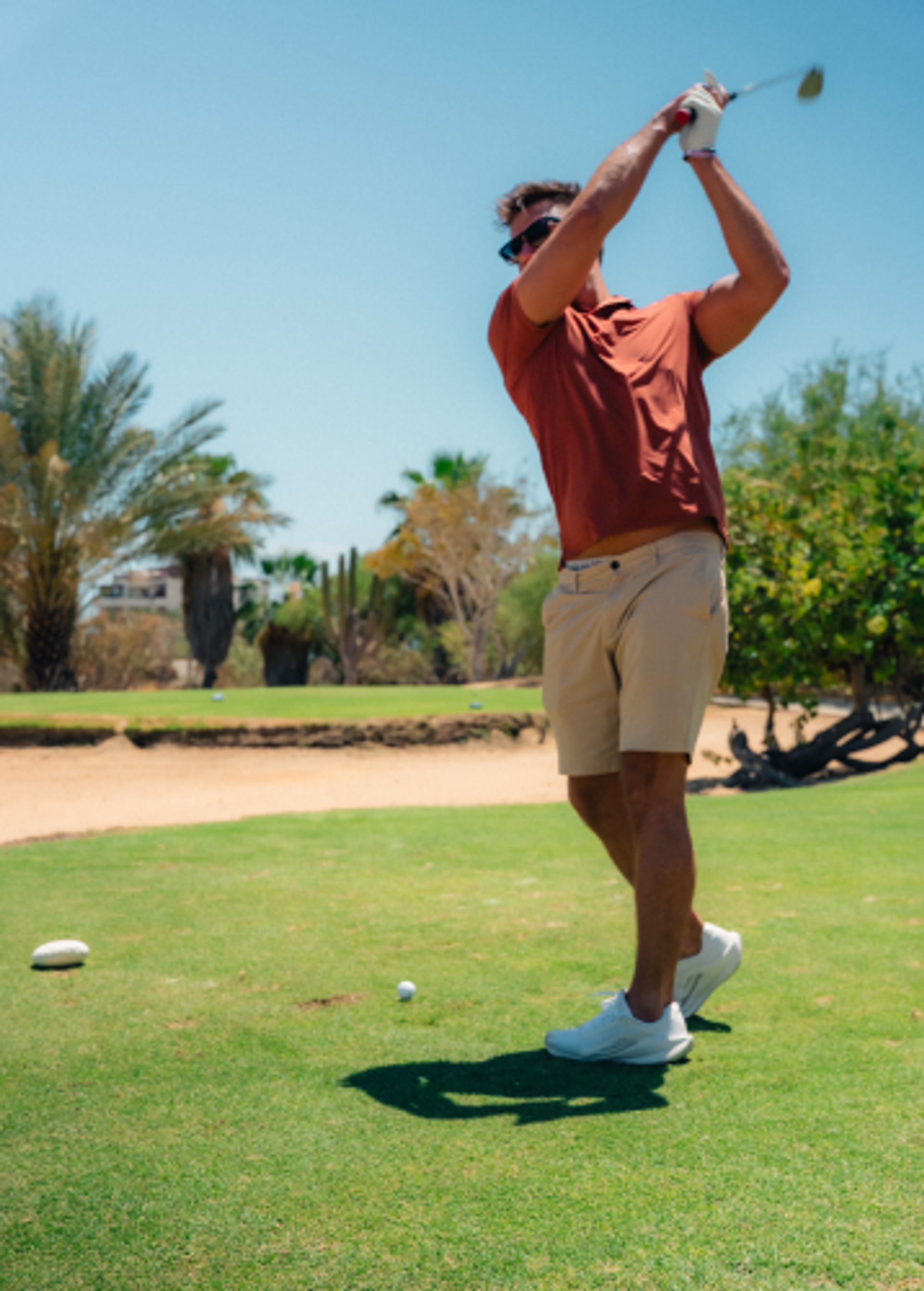 A man in sunglasses and a rust-colored polo shirt swings a golf club on a sunny golf course.