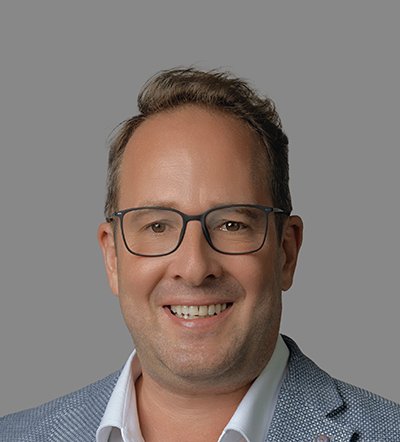 Headshot of a smiling man wearing glasses and a light-colored blazer against a gray background.