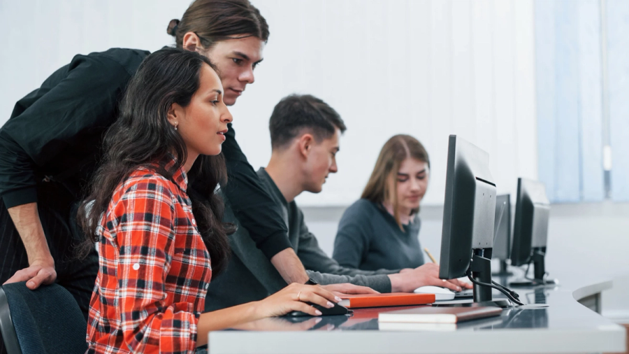 Four students in a computer lab, one assisting another at a desktop computer.