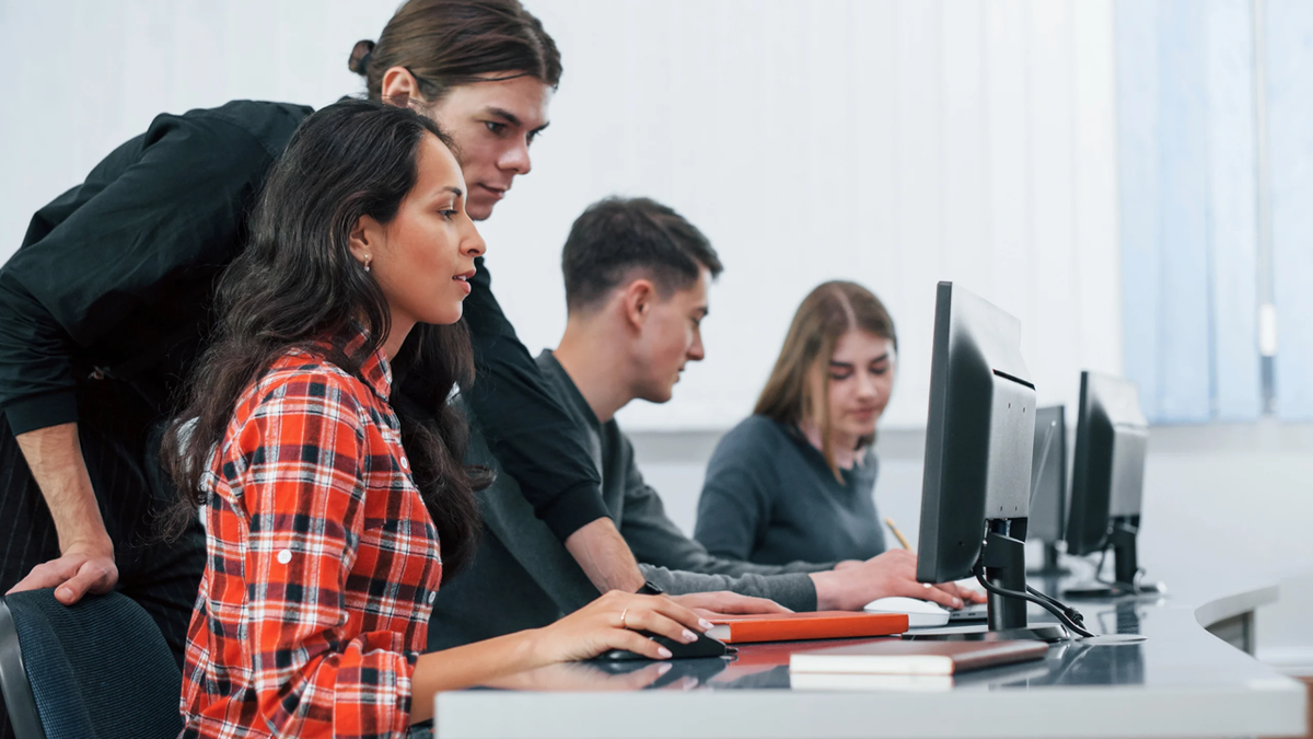 Four students in a computer lab, one assisting another at a desktop computer.