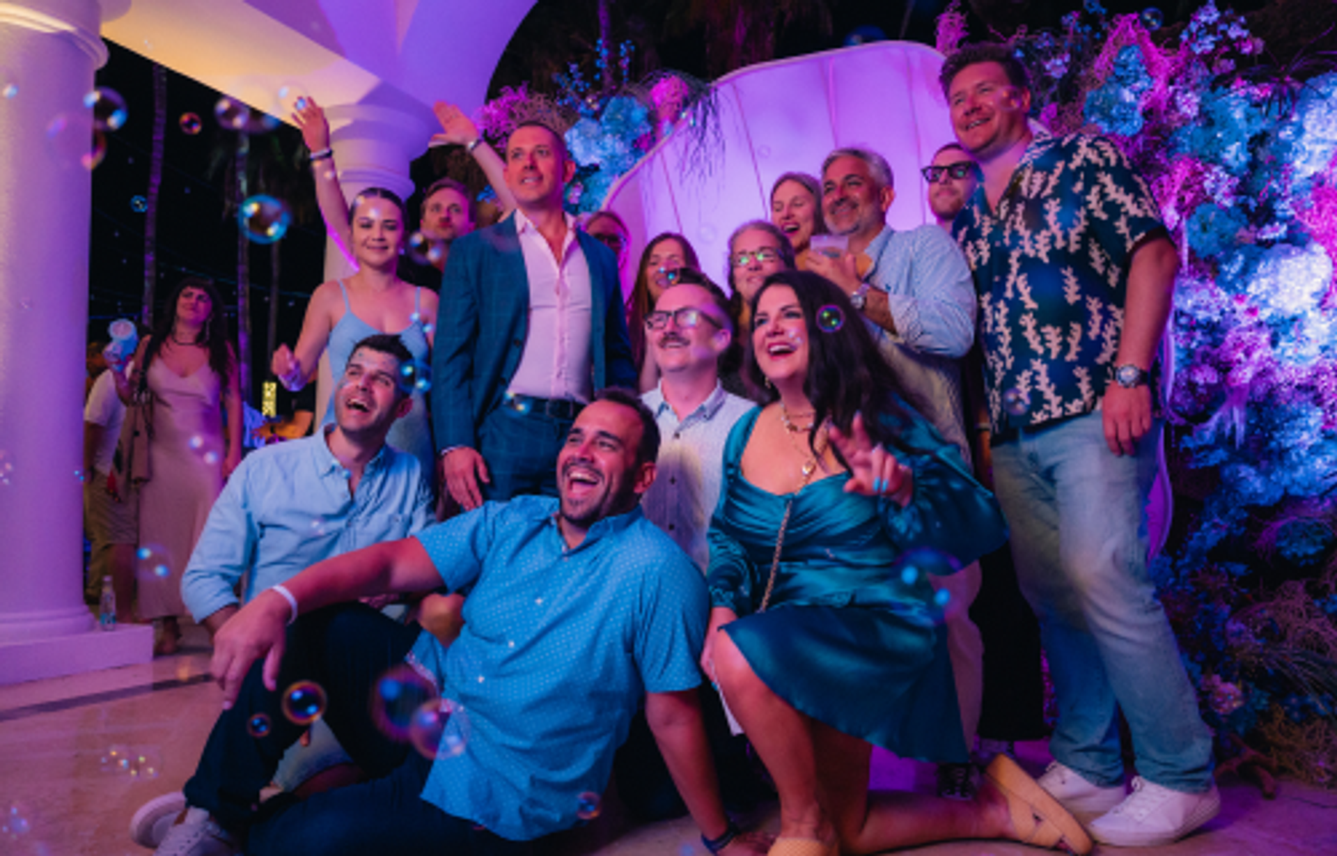 A group of happy people posing for a photo at a vibrant evening event with bubbles and purple and blue lighting.