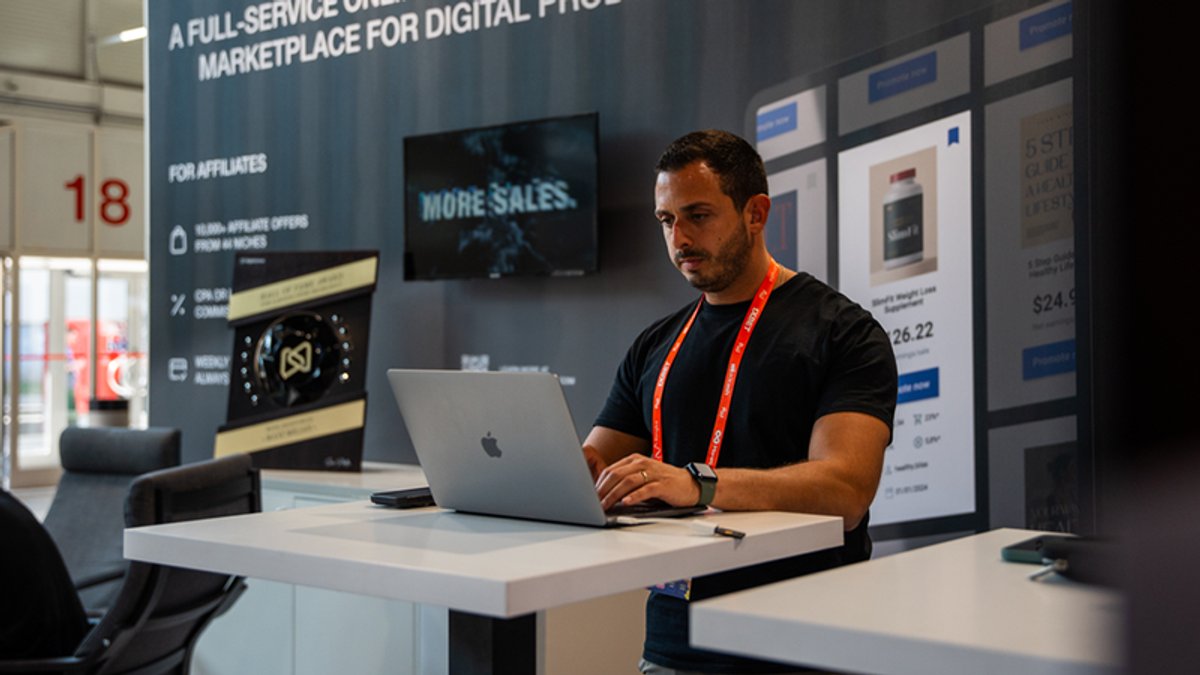 A man works on a laptop at an exhibition booth with marketing displays.