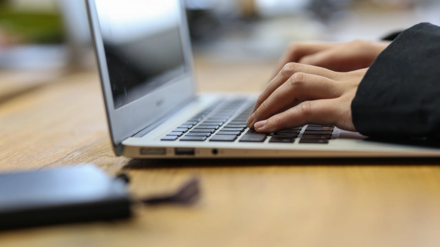 Close-up of hands typing on a silver laptop on a wooden desk.