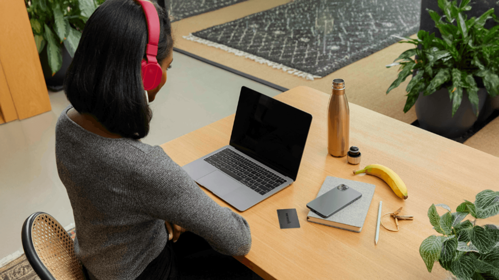A person wearing red headphones sits at a wooden desk with a laptop, gold water bottle, and a banana.