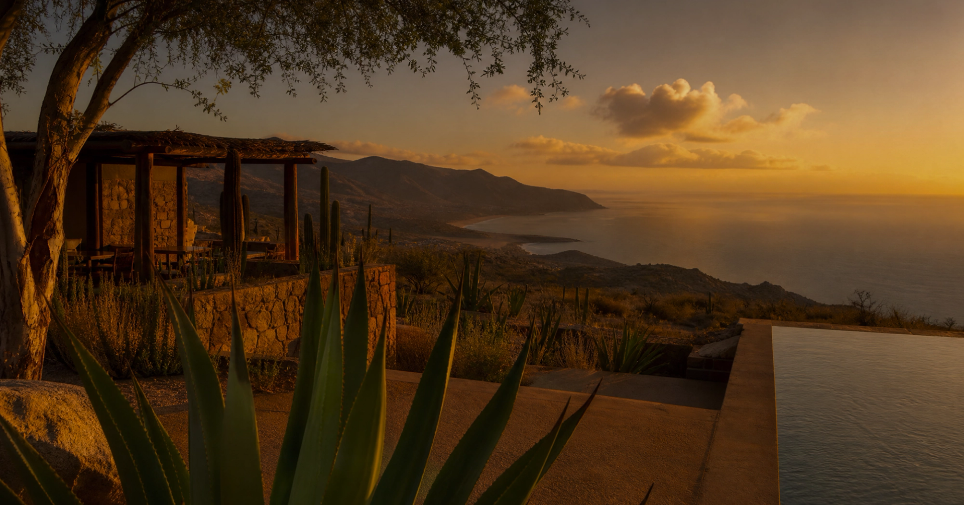 A golden sunset over a mountainous coast and ocean, viewed from a patio with an infinity pool.