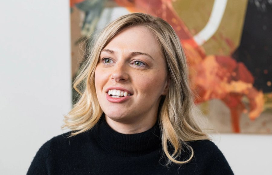 A blonde woman in a black turtleneck smiles, with an abstract painting in the background.