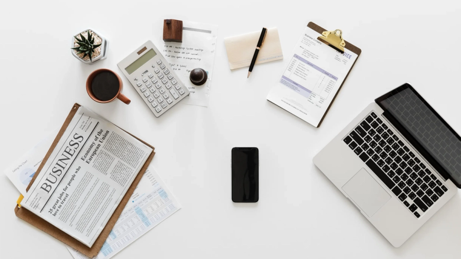 Overhead view of a white business desk with a laptop, newspaper, calculator, coffee, and smartphone.