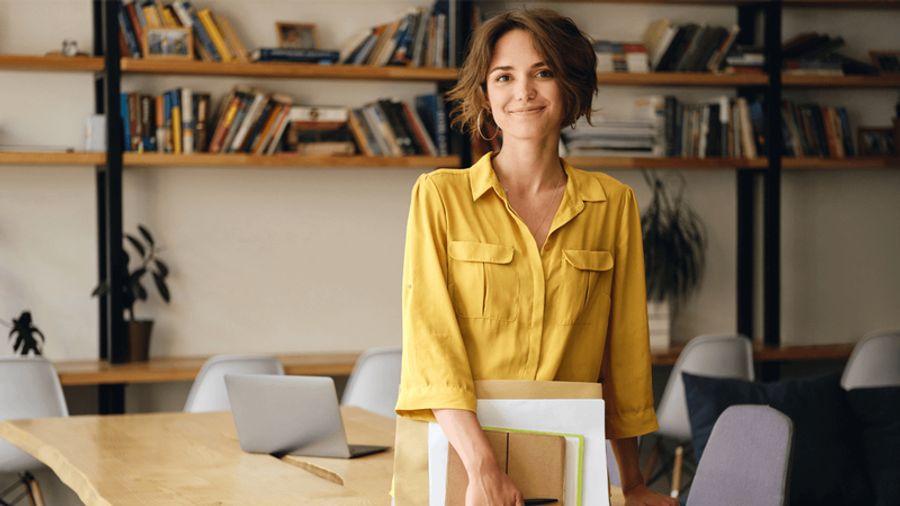 Smiling woman in a yellow shirt holding documents in a modern office.