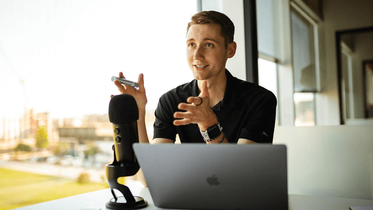 A young man speaking into a microphone at a desk with a laptop.