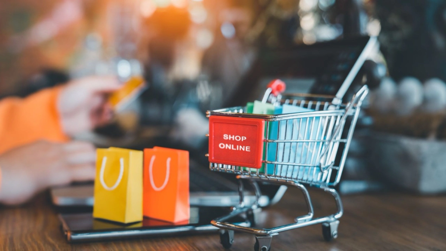 Miniature shopping cart with "SHOP ONLINE" sign, shopping bags, and a person using a credit card on a laptop.