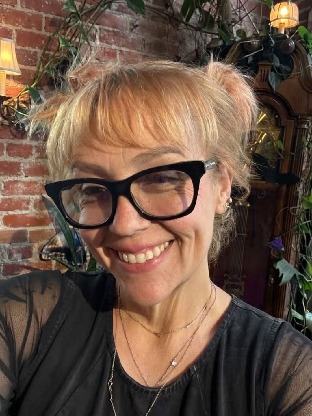 A smiling woman with blonde pigtails and black glasses wears a black top and layered necklaces against a brick wall with plants.