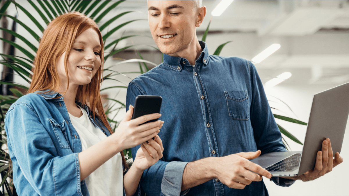 A smiling woman shows a man something on her smartphone while he holds a laptop.
