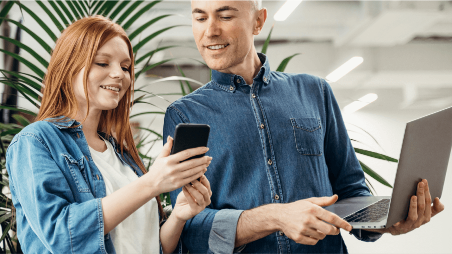 A smiling woman shows a man something on her smartphone while he holds a laptop.