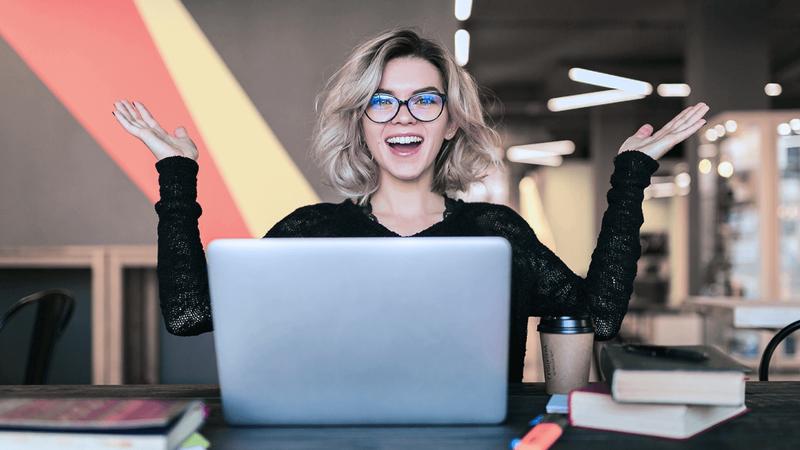 A smiling woman with glasses and blonde hair raises her hands in excitement behind a laptop.