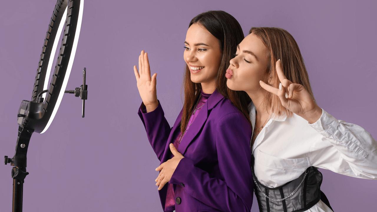 Two young women pose for a video with a ring light and smartphone.