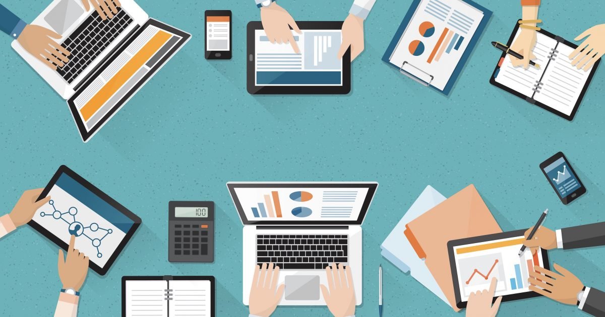 Overhead view of multiple hands working on laptops, tablets, and documents at a collaborative workspace.