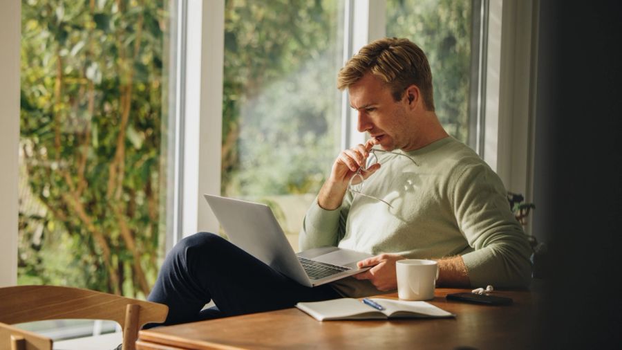 Thoughtful man working on a laptop at a table by a window.
