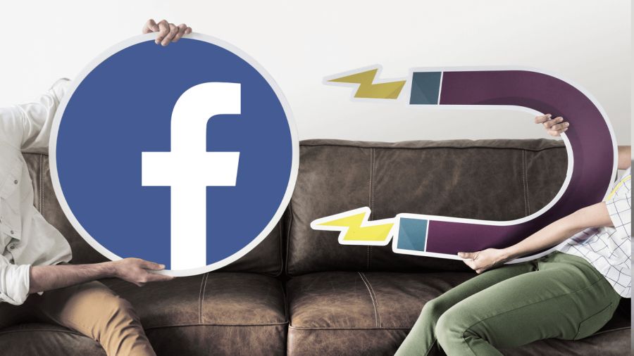 A person holds a large Facebook logo sign opposite another person holding a large magnet sign, seated on a brown couch.