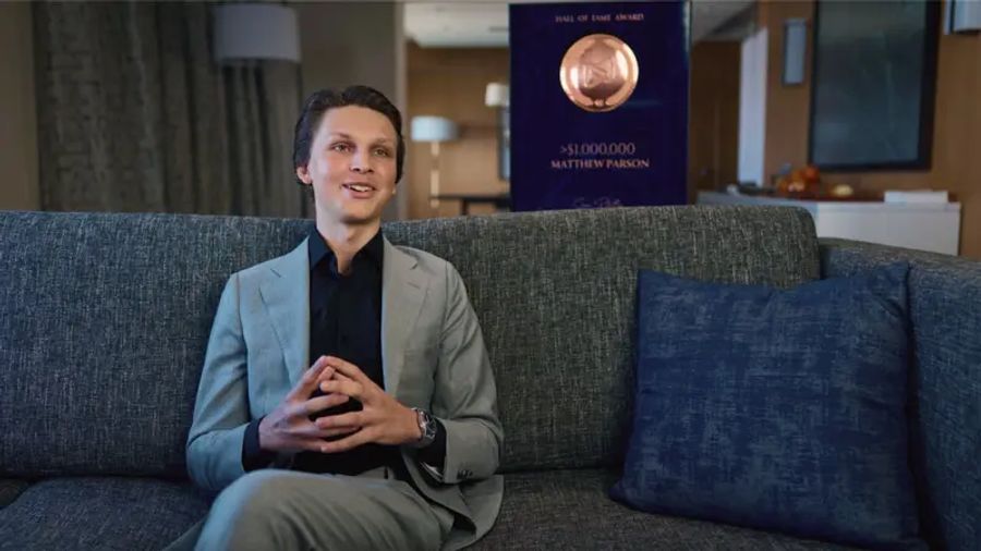 A smiling man in a suit sits on a gray couch, with a "Hall of Fame Award" plaque for "Matthew Parson" and ">$1,000,000" visible behind him.