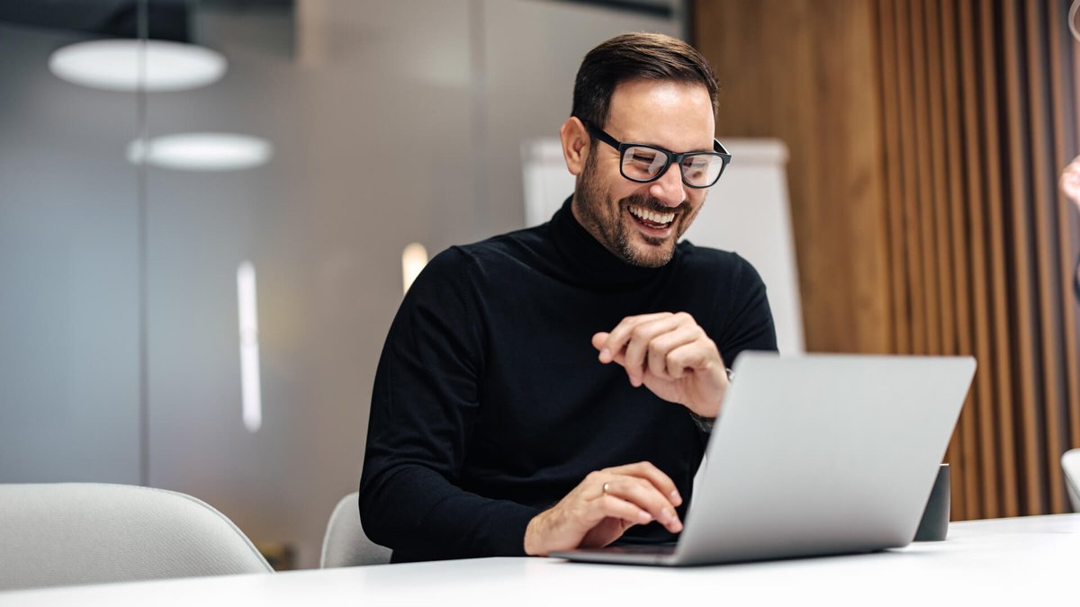 Man in a black turtleneck and glasses laughing at his laptop.