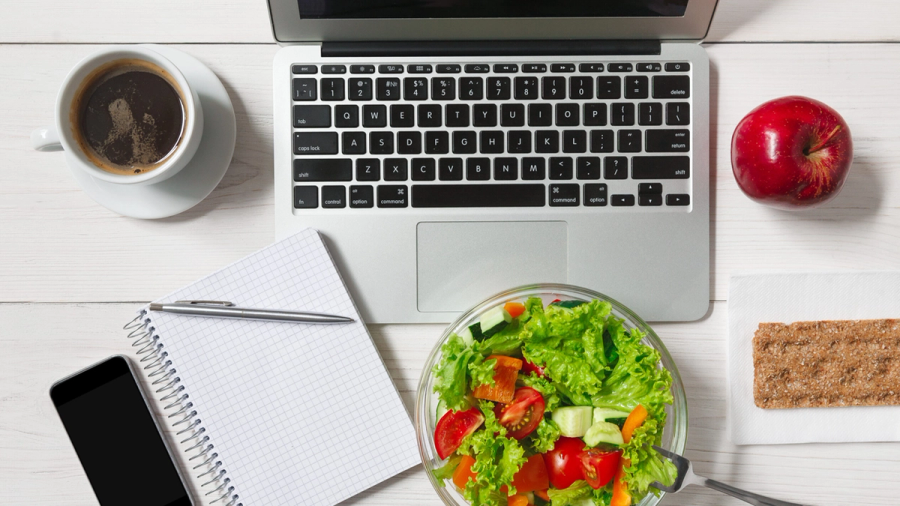 Overhead view of a laptop, coffee, salad, apple, crispbread, notepad, and smartphone on a white desk.