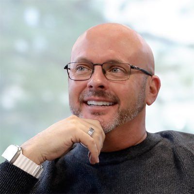 A smiling bald man with a beard and glasses rests his chin on his hand while looking up.