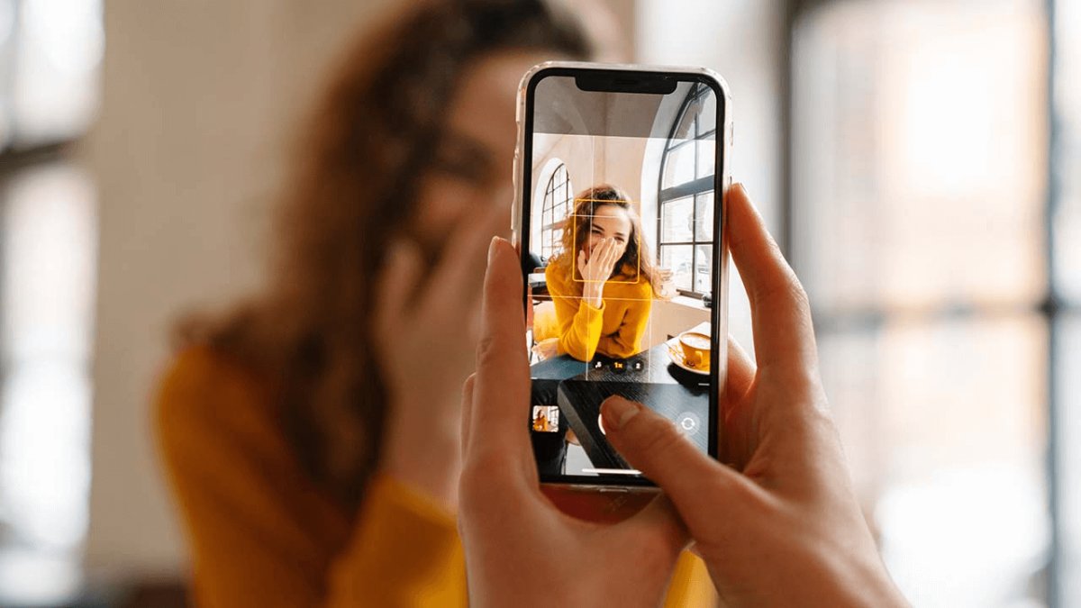 Hands holding a smartphone, taking a photo of a smiling woman covering her mouth.
