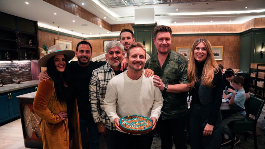 A group of eight smiling people posing with a man holding a large round cookie cake in a lounge area.