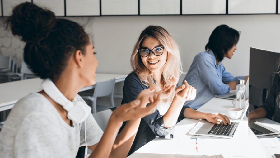 Two women smiling and talking at desks in a bright office.