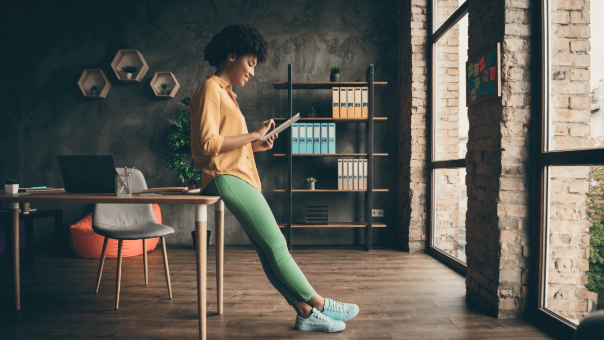 A Black woman in an office leans on a desk while looking at a tablet.