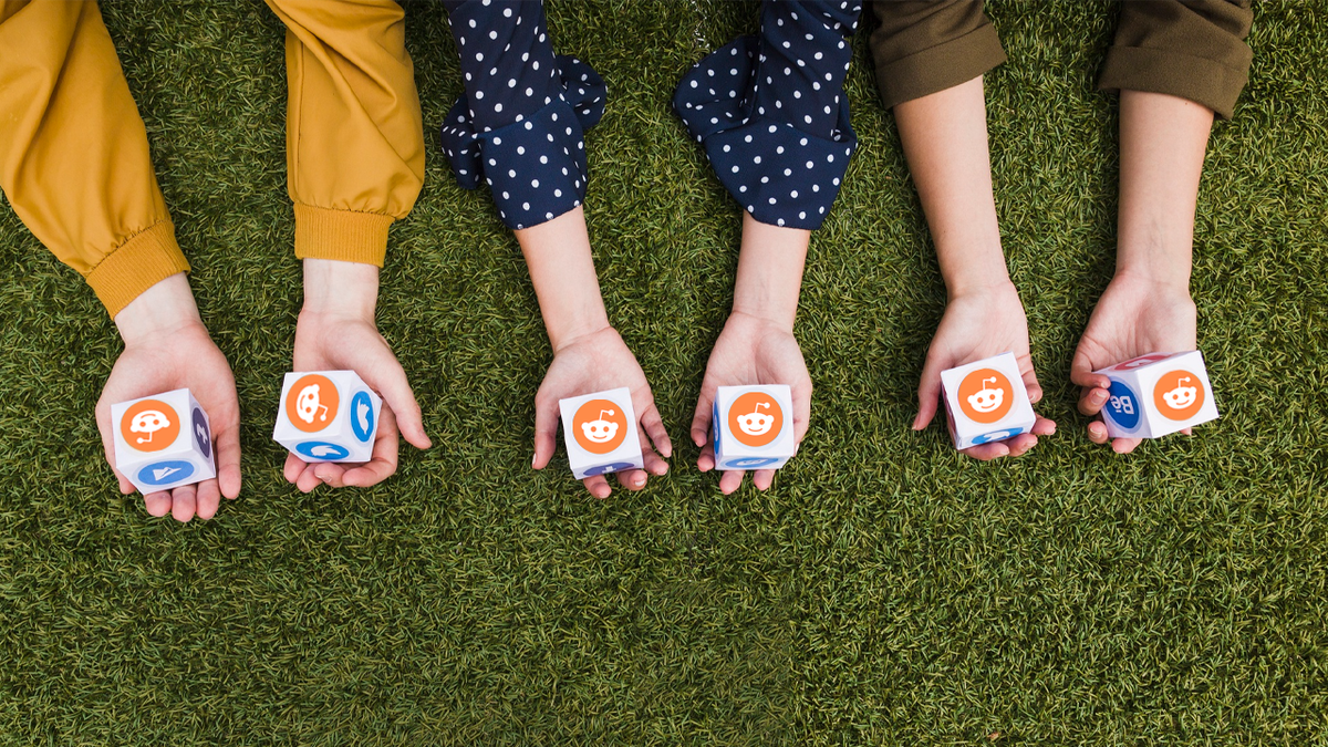 Four hands holding white cubes with orange Reddit logos on a green surface.