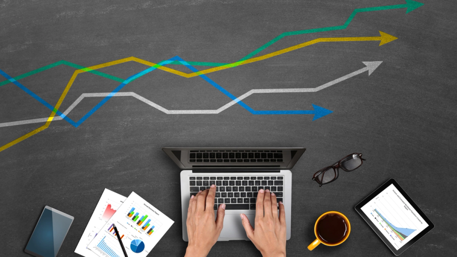 Hands typing on a laptop at a desk with business charts and upward-pointing trend lines.