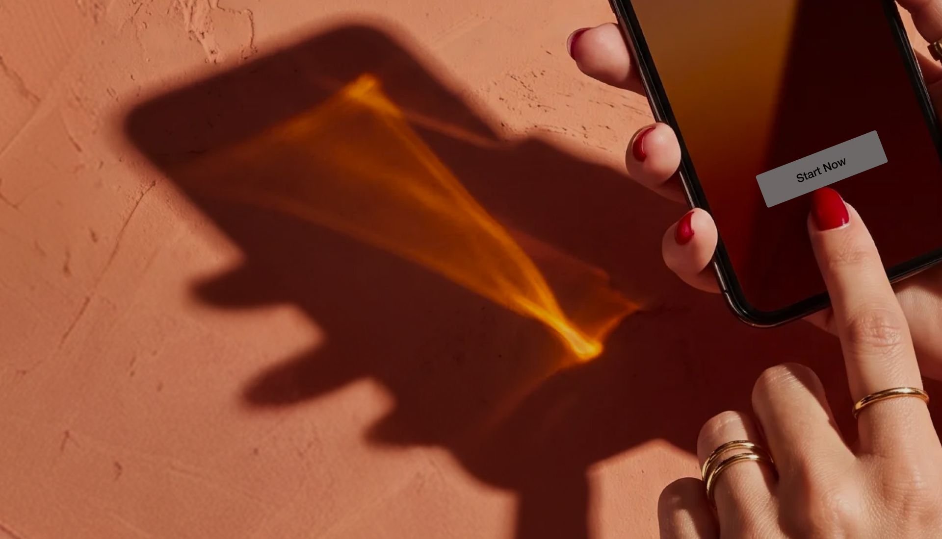 Hand tapping "Start Now" on a smartphone, with light refraction and shadows on a textured terracotta background.
