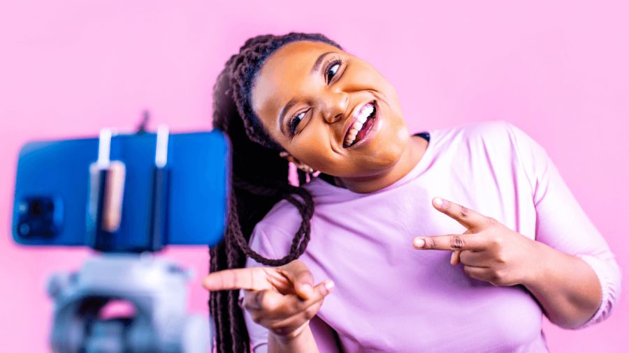 A smiling Black woman with braids records a video on a smartphone mounted on a tripod, pointing at the camera, against a pink background.