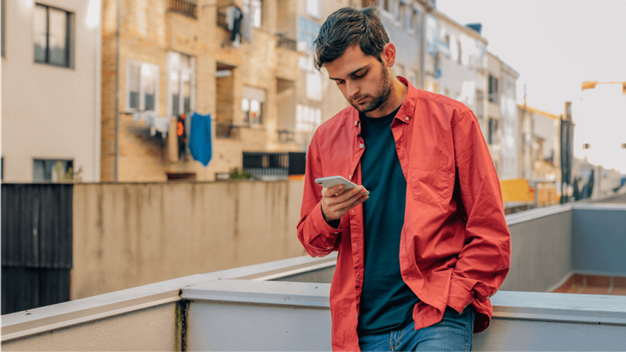 A young man in a red shirt intently looks at his phone on a balcony.