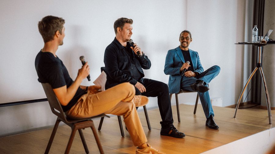 Three men on a stage, seated and holding microphones for a panel discussion.