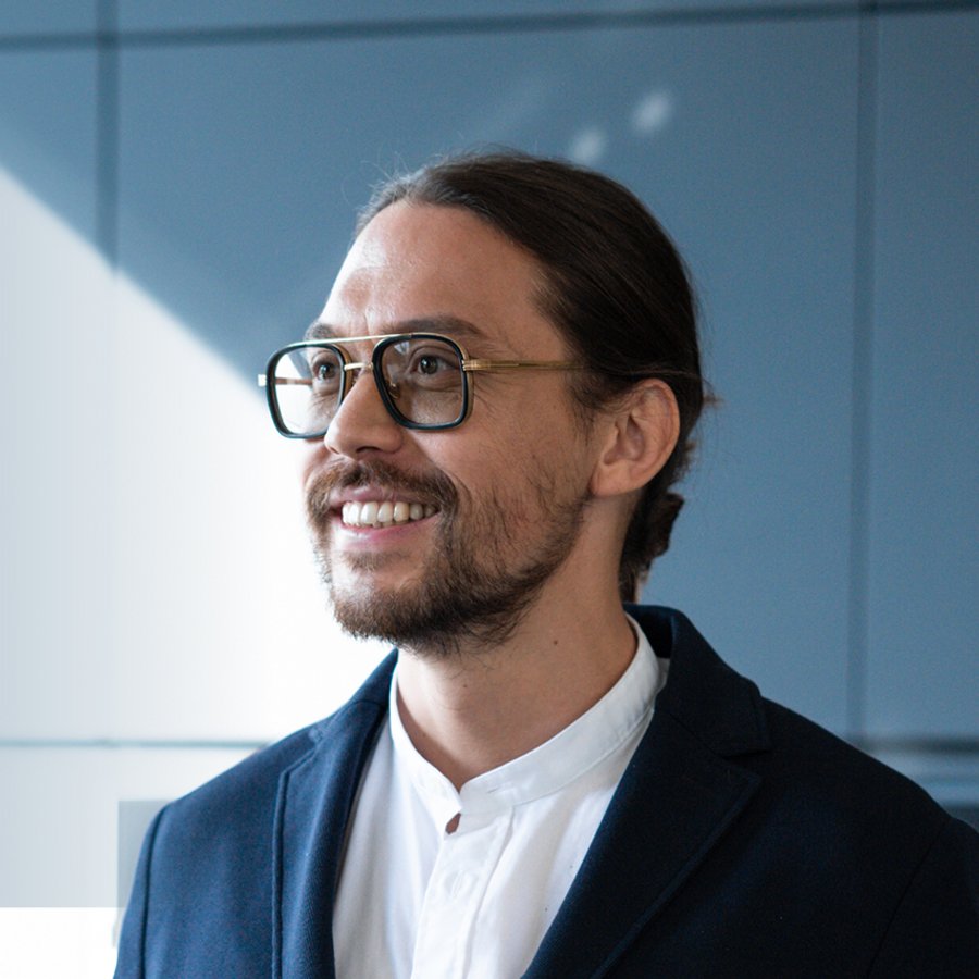 Hombre sonriente con cabello largo recogido, barba y gafas, con chaqueta oscura sobre camisa blanca.