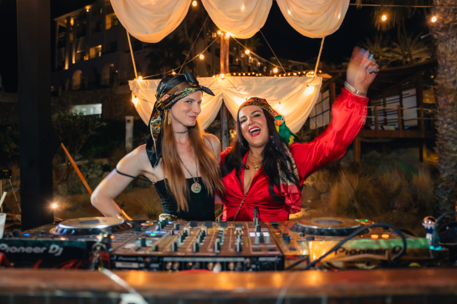Two women in pirate costumes behind a DJ mixer, one smiling with a parrot on her shoulder, at an outdoor night party.