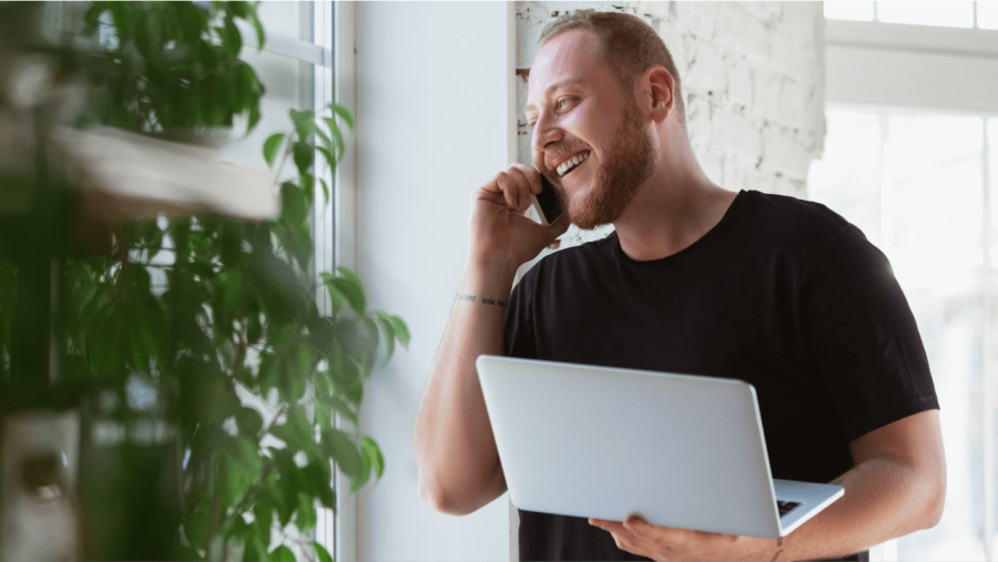 A smiling man with a beard talks on a phone and holds a laptop.