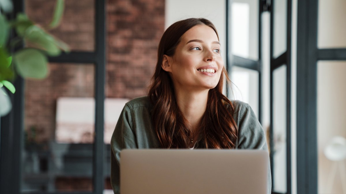 A smiling young woman looks to the right from behind a laptop.