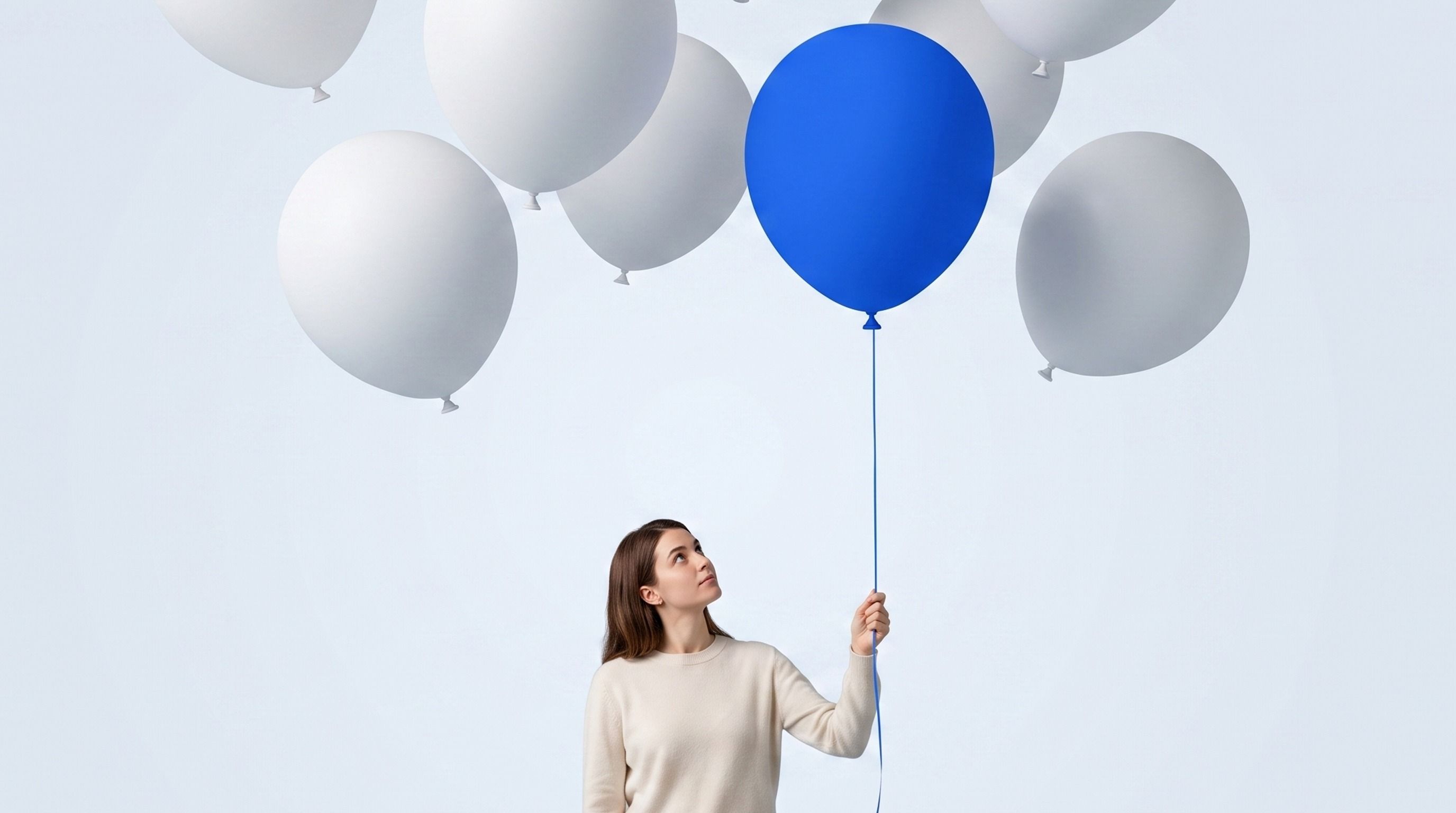 A woman holds a blue balloon while looking up at several white balloons floating above her.