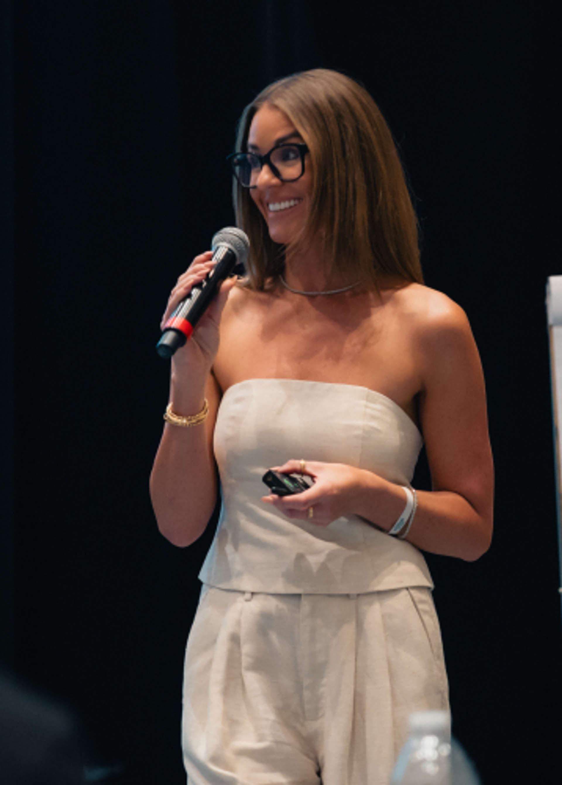 A smiling woman with glasses, wearing a light-colored strapless outfit, holds a microphone and a remote while presenting.
