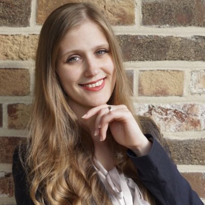 A smiling woman with long blonde hair, red lipstick, and a blazer, resting her chin on her hand, against a brick wall.